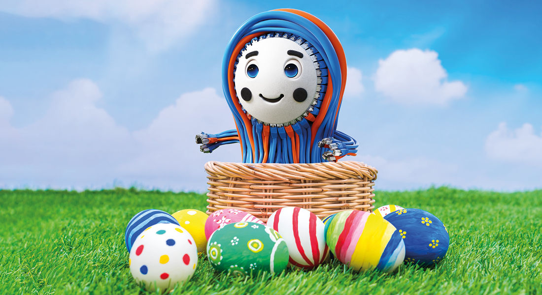 Colorful Easter eggs and Patchy, the cheerful mascot of EFB-Elektronik, in a basket on green grass in front of a blue sky.