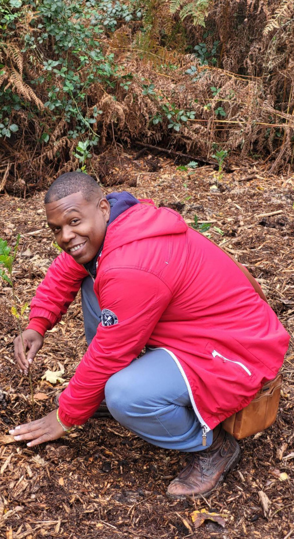 Mann in roter Outdoor-Jacke pflanzt lächelnd einen Setzling bei einer Baumpflanzaktion im Wald