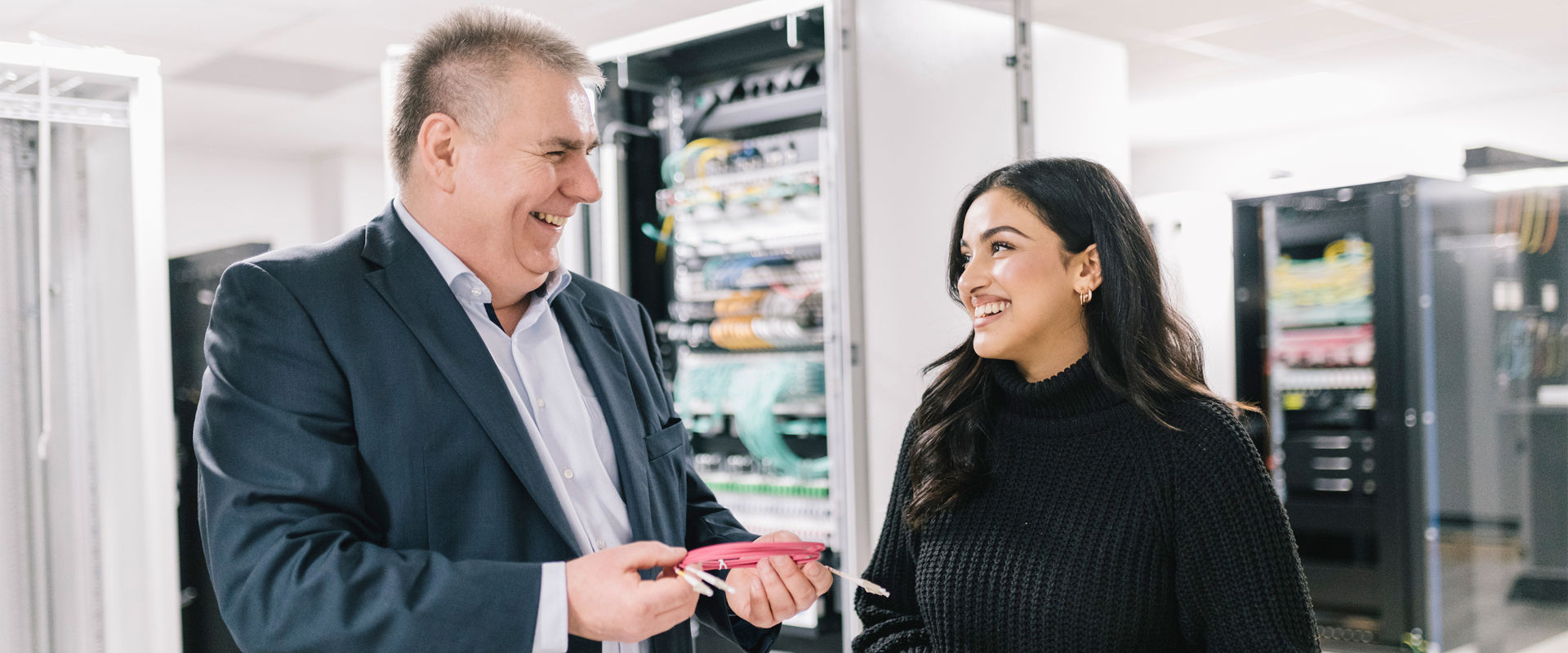 Two smiling people in the server room, man holding a pink fiber optic patch cable.