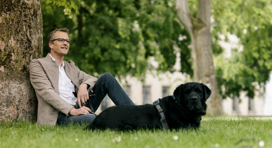 Mann sitzt an einem Baum lehnend auf einer Wiese. Neben ihm sitzt ein großer, schwarzer Hund