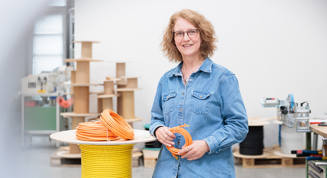 Frau in Jeanshemd hält eine Rolle orangefarbenes Kabel, große Kabeltrommel mit gelbem Kabel im Vordergrund, Kabeltrommeln und Geräte im Hintergrund.