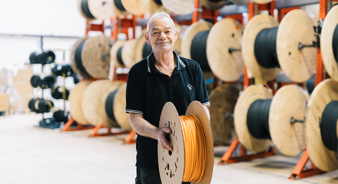 Man holding a reel of orange cable, several large cable drums in the background.