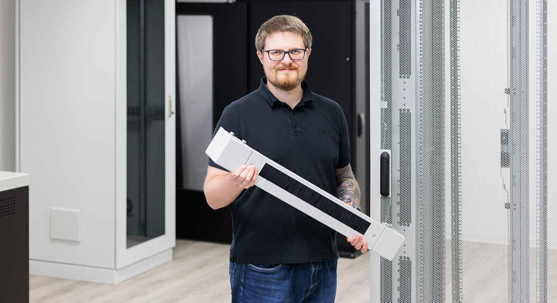 Man in black polo shirt holding an elongated white component with brushes, standing in front of server or network cabinets