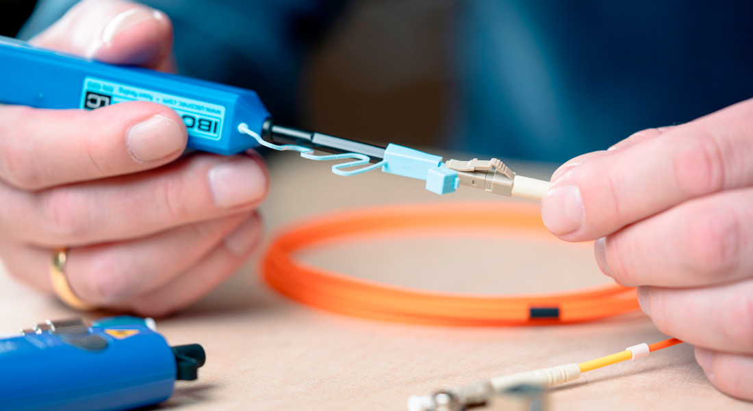Close-up of a person cleaning a fiber optic cable with a cleaning tool, in the background more fiber optic cables and an adapter.